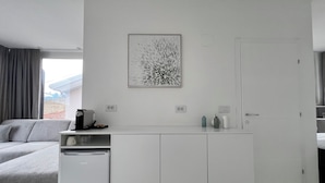 A modern, minimalist room with a grey sofa, a white kitchenette, and a large window with curtains.