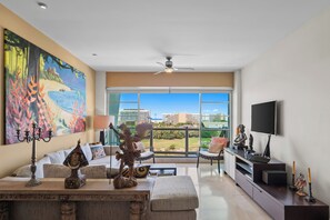 A modern living room with a large window, a ceiling fan, and a flat-screen TV. - Luxury Beach Condo with Resort Access Vidanta (Nuevo Vallarta)