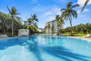 Uma piscina com espreguiçadeiras, palmeiras e um prédio de hotel ao fundo.