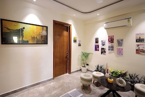 A modern interior with a wooden door, a framed artwork, a potted plant, a round table with stools, and a wall-mounted air conditioner. - ZEALIN Stays Golf Course Road PHASE 1 (Gurugram)