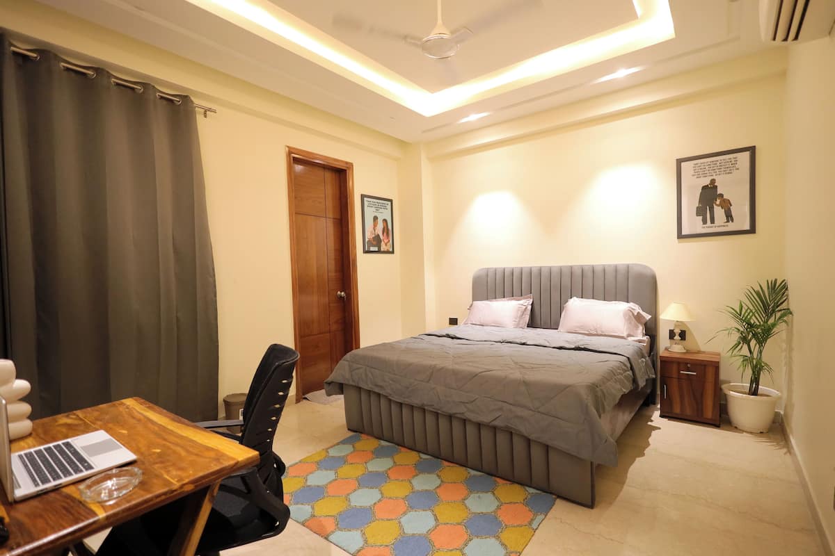 Quarto com cama, escrivaninha com laptop, cadeira, mesa de cabeceira com abajur e planta em vaso.