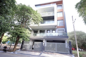 Un edificio residencial de varios pisos con entrada protegida, un balcón en el nivel superior y una zona de estacionamiento.