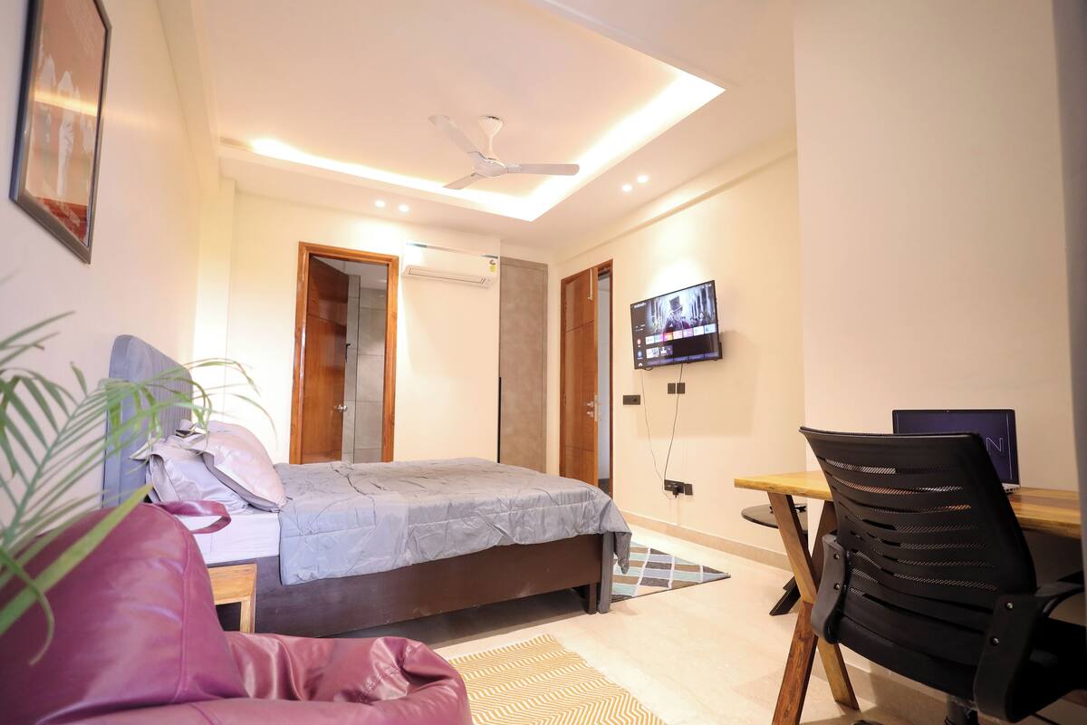 Quarto com cama, escrivaninha, cadeira, televisão e ventoinha no teto.