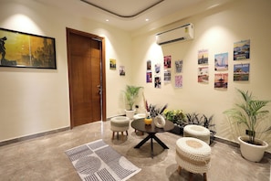 A well-lit hallway with a wooden door, a round table with stools, potted plants, and wall posters of various cities. - ZEALIN Stays Golf Course Road PHASE 1 (Gurugram)