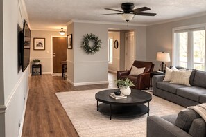 Ein Wohnzimmer mit Deckenventilator, Kamin, grauem Sofa, Couchtisch und einem Essbereich mit Holztisch und Stühlen.