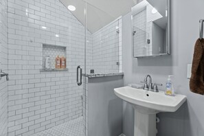 A bathroom with white subway tiles, a shower with a glass door, a sink with a mirror, and toiletries on the counter. - Entire Midtown 4plex - Gated Parking - Rail to NRG (Houston)