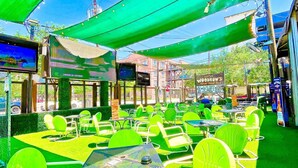 An outdoor seating area with green chairs and tables, a TV screen, and a sign for 'Little Woodrow's.' - Entire Midtown 4plex - Gated Parking - Rail to NRG (Houston)