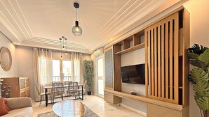 Ein Wohnzimmer mit einer Couch, einem Esstisch mit Stühlen, einem Fernseher auf einem Holzständer, einer großen Topfpflanze und einer Deckenleuchte.