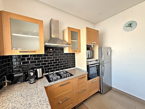 Une cuisine moderne avec des meubles en bois, un pare-feu en carreaux noirs, des appareils en acier inoxydable et une horloge au mur.
