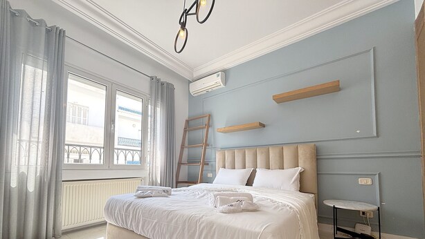 Ein Schlafzimmer mit einem großen Bett, einem Heizkörper, einem Fenster mit Vorhängen, einer Stehleiter, einem Nachttisch und einem an der Wand befestigten Regal.