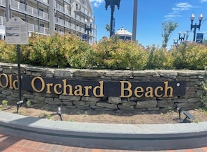 A stone wall with 'Old Orchard Beach' sign, a building in the background, and a clear sky.