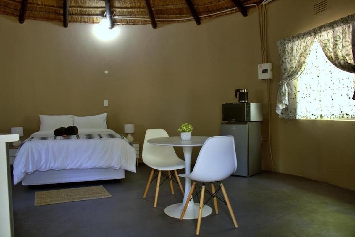 Un dormitorio con cama, una mesita con sillas, cafetera y ventana con cortinas.