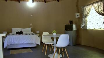 Un dormitorio con cama, una mesita con sillas, cafetera y ventana con cortinas.