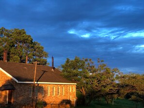 A brick building with a sloped roof, multiple chimneys, and a thatched roof structure to the left. - Rising Rocks Farm (Magaliesburg)