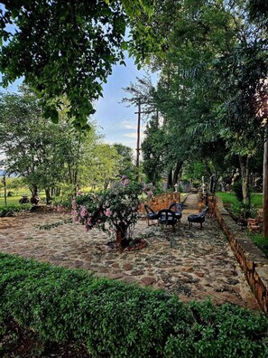 A stone patio with a seating area, surrounded by lush greenery and trees.