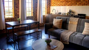 A room with a brick wall, a sofa, a table, and a kitchen area. - Rising Rocks Farm (Magaliesburg)