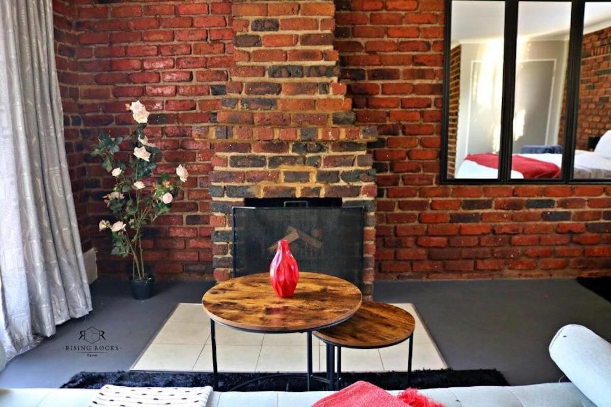 Une pièce avec une cheminée en briques, une table ronde en bois surmontée d’un vase rouge et un canapé.
