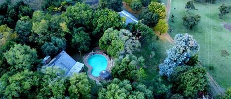 An aerial view of a property with a swimming pool surrounded by dense trees, a visible road, and a fenced area.