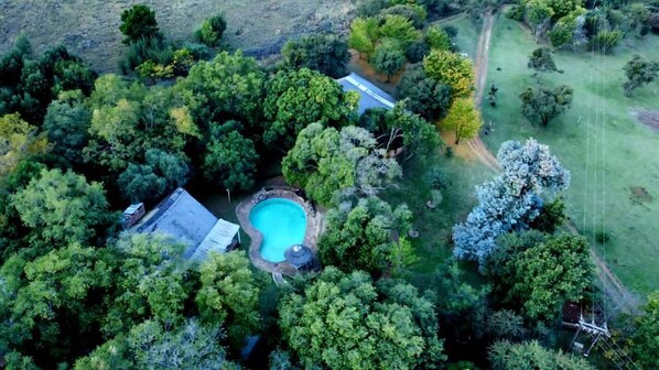 An aerial view of a property with a swimming pool surrounded by dense trees, a visible road, and a fenced area.