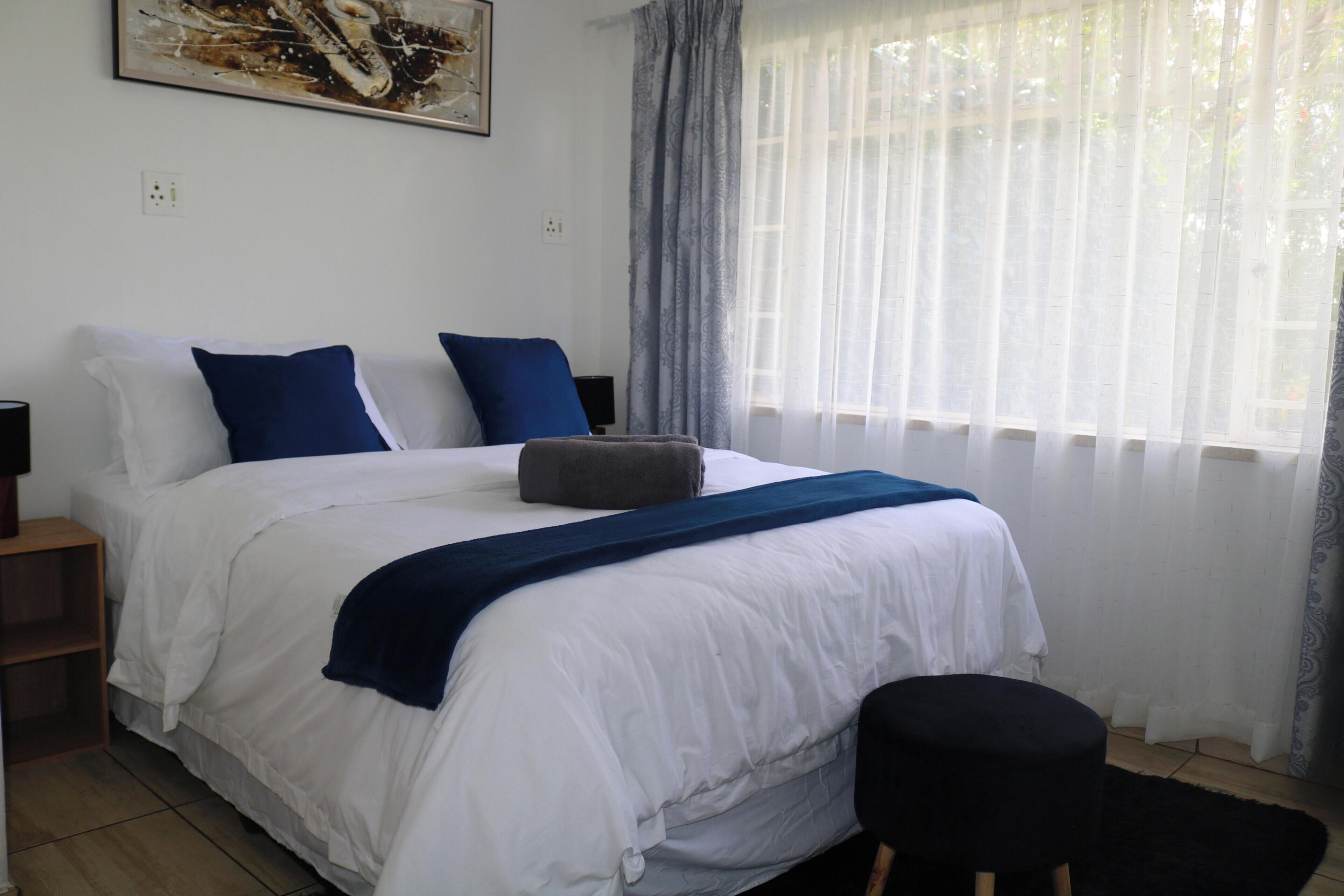A bedroom with a bed, pillows, a bedside table, a lamp, a window with curtains, and a painting on the wall.
