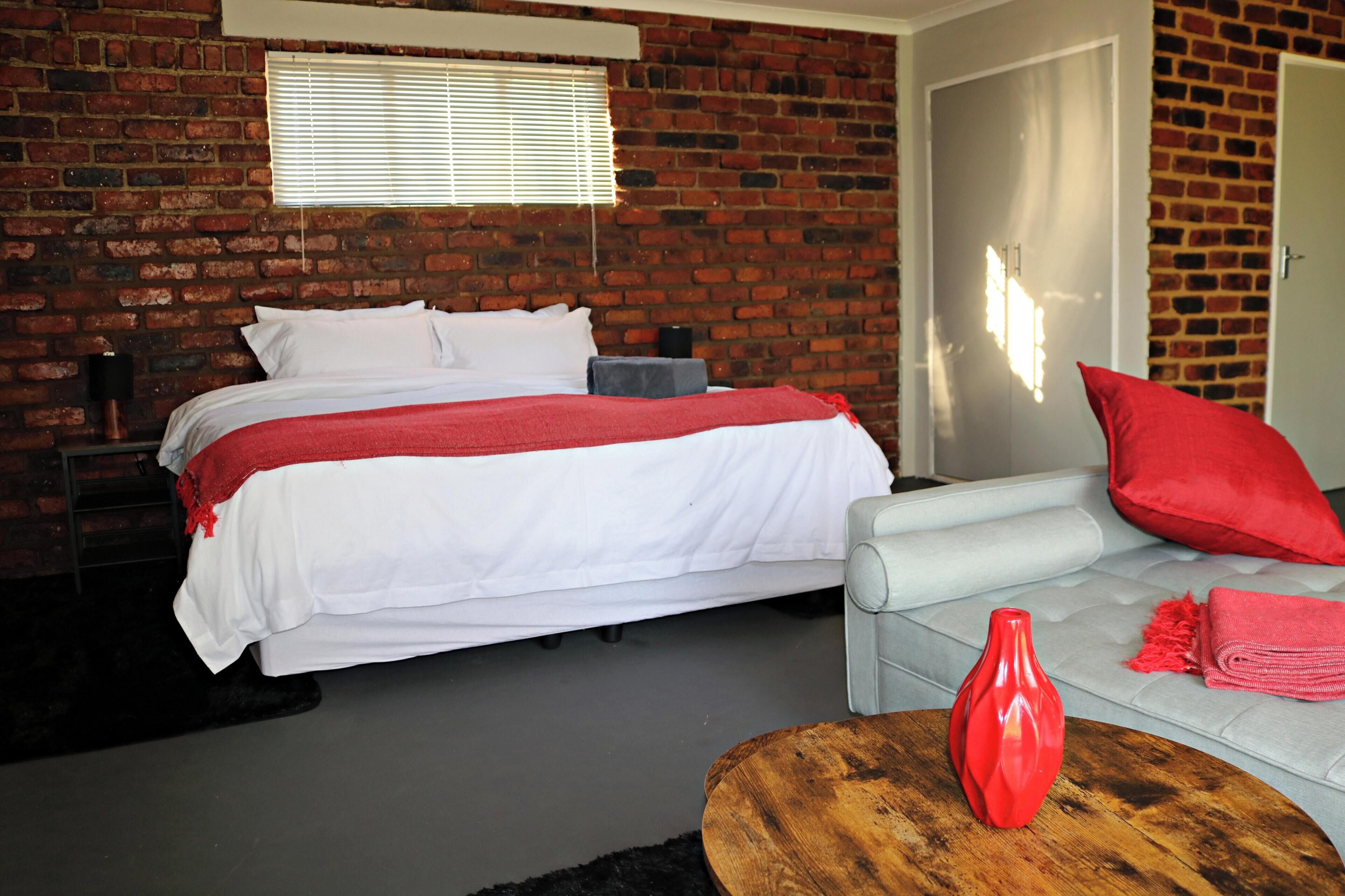 A bedroom with a brick wall, a bed with white bedding, a grey sofa with red cushions, and a wooden coffee table with a red vase.