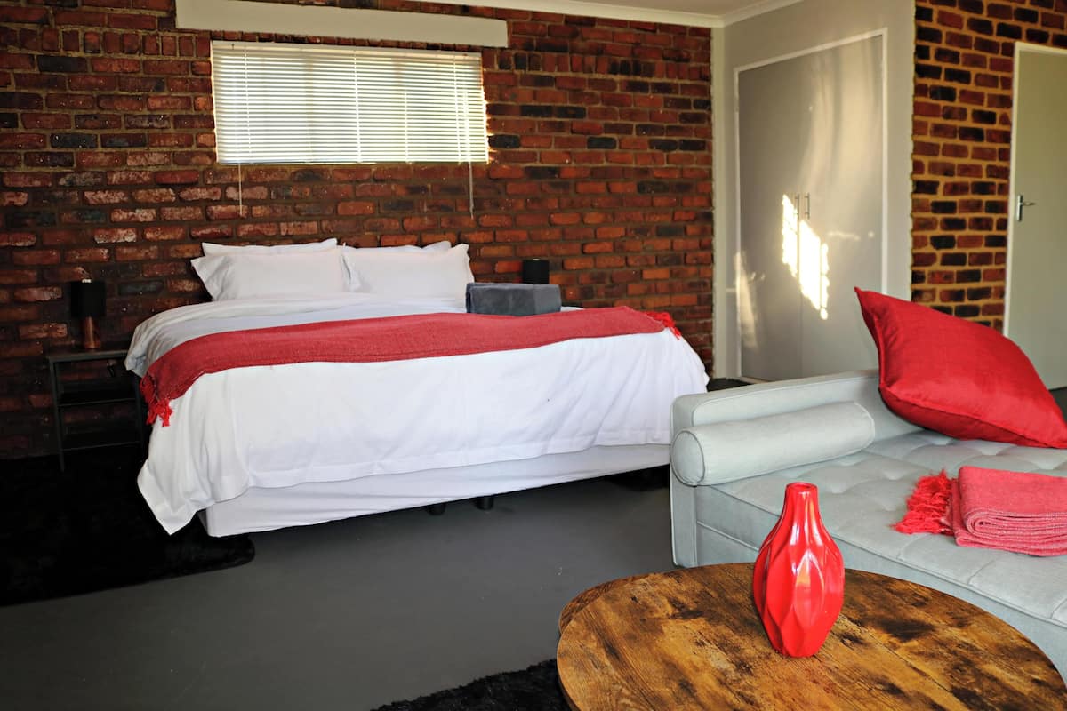 A bedroom with a brick wall, a bed with white bedding, a grey sofa with red cushions, and a wooden coffee table with a red vase.