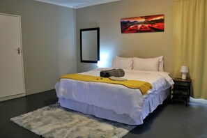 A hotel room with a bed, a nightstand, a lamp, a painting on the wall, and a door. - Rising Rocks Farm (Magaliesburg)