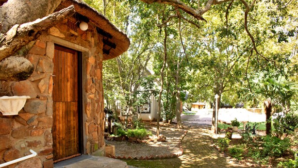 A stone cottage with a wooden door, a stone staircase, and a garden with trees and a paved path. - Rising Rocks Farm (Magaliesburg)