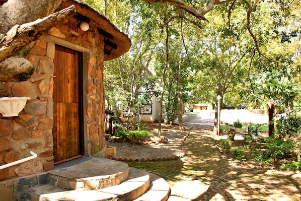 Una caseta de pedra amb una porta de fusta, una escala de pedra i un jardí ple d'arbres i un camí pavimentat.