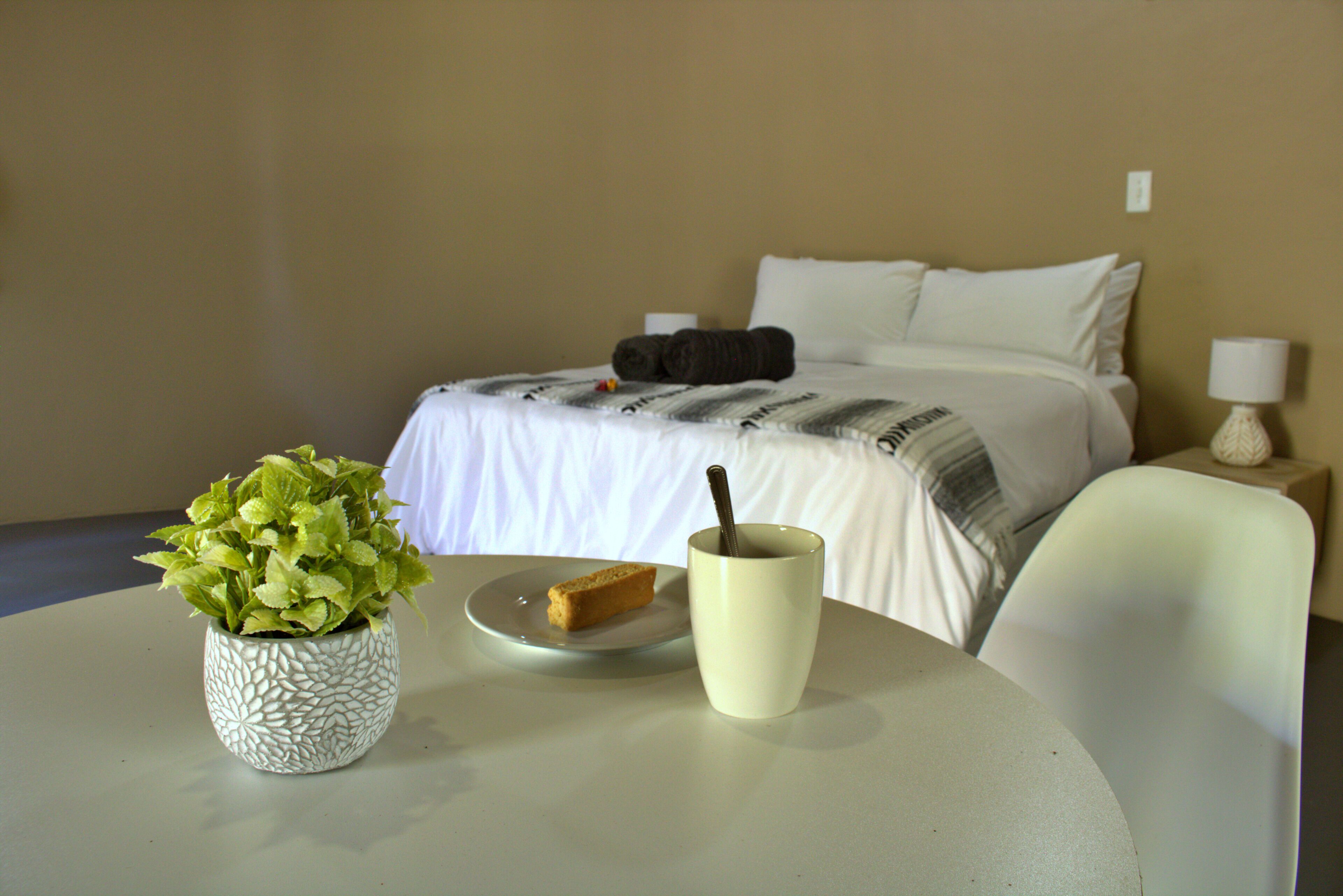 A hotel room with a bed, a bedside table with a lamp, a small table with a plant and a plate with toast, and a cup with a spoon.