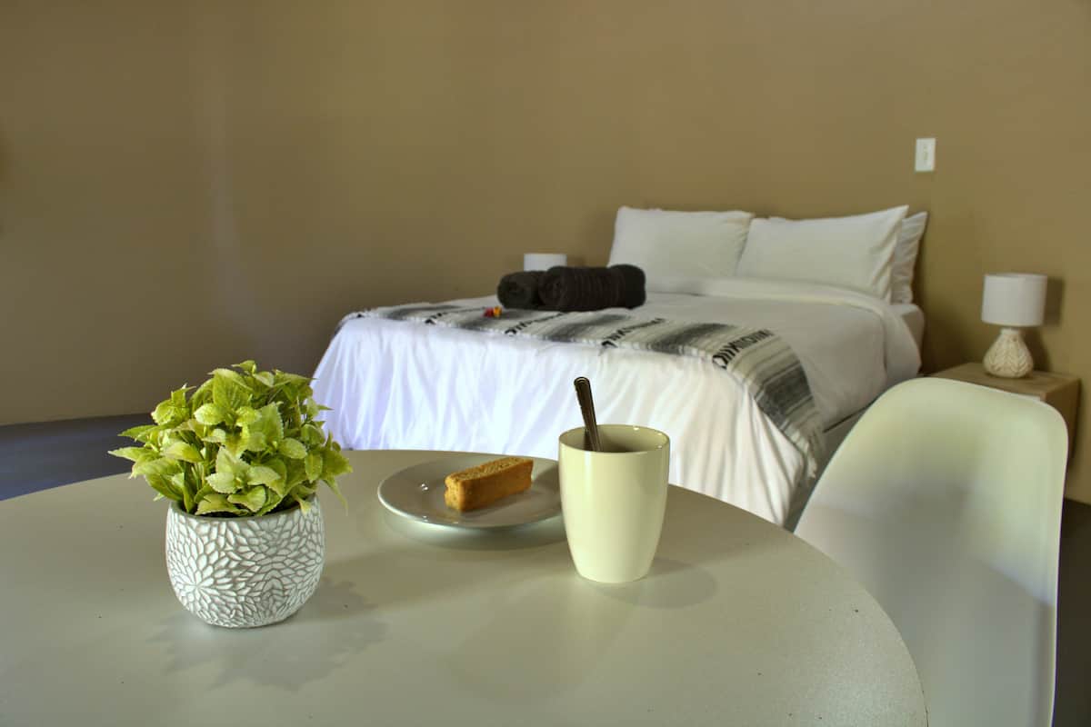 A hotel room with a bed, a bedside table with a lamp, a small table with a plant and a plate with toast, and a cup with a spoon.