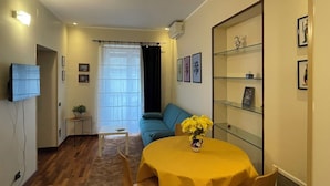 Ein Wohnzimmer mit einer Couch, einem Fernseher, einem Esstisch mit gelber Tischdecke und einer Vitrine mit Bildern.