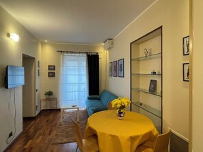 Ein Wohnzimmer mit einer Couch, einem Fernseher, einem Esstisch mit gelber Tischdecke und einer Vitrine mit Bildern.