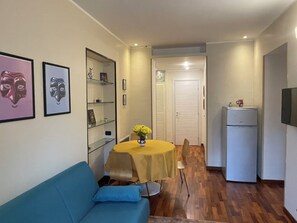 Un espai vital compacte amb un sofà, una taula de dinar amb un ram de flors, un frigorífic i una porta que dona a una altra estança.