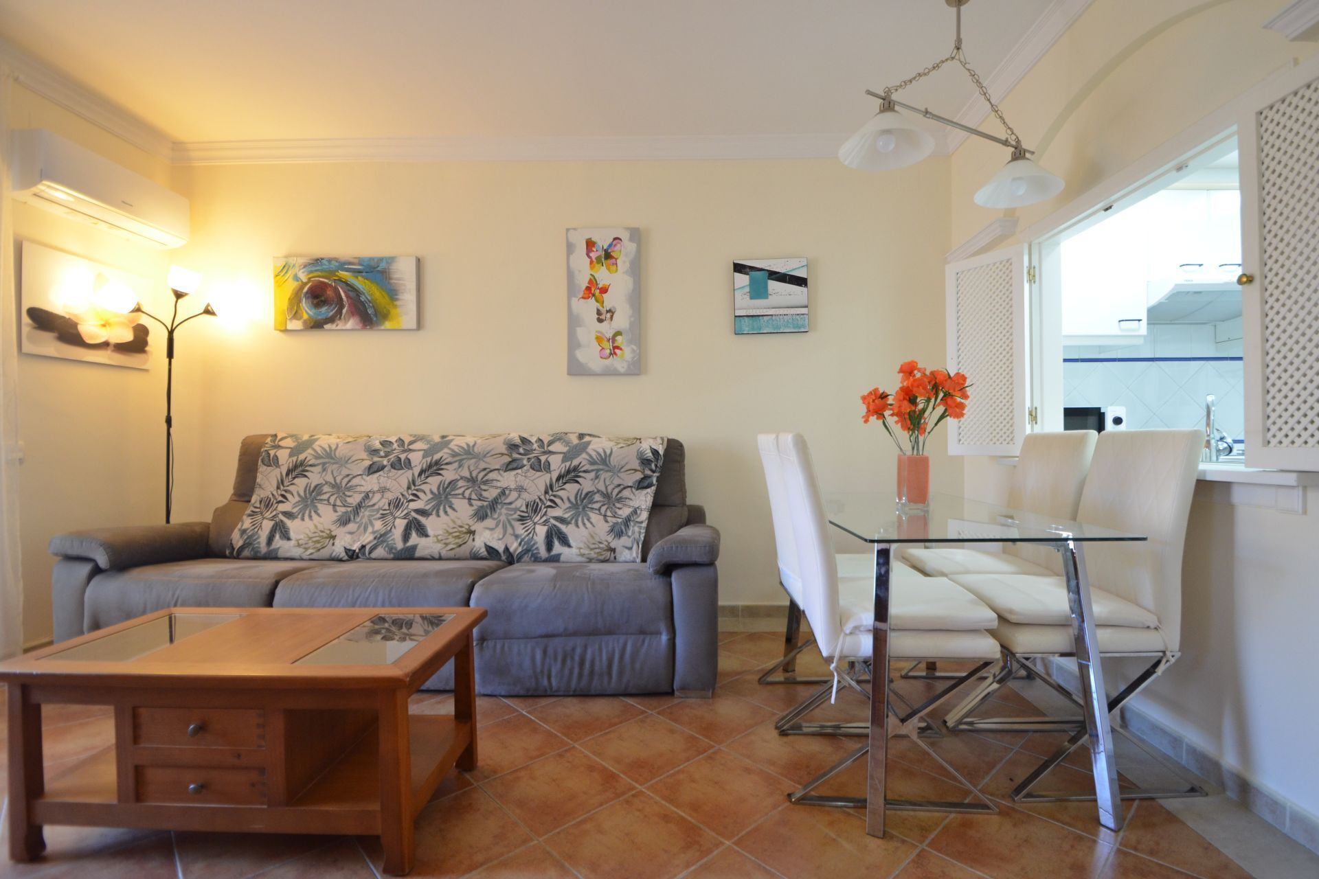 A living room with a sofa, a glass table, and a chair. There are paintings on the wall and a window with blinds.
