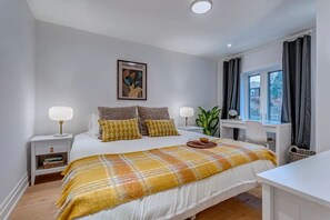 Une chambre à coucher comprenant un lit, des tables de chevet, un bureau, une chaise, une fenêtre avec des rideaux et une plante.
