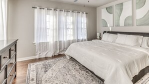 Ein Schlafzimmer mit Bett, Deckenventilator, Nachttisch, Kommode und Fenster mit Vorhängen.