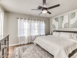 Ein Schlafzimmer mit Bett, Deckenventilator, Nachttisch, Kommode und Fenster mit Vorhängen.