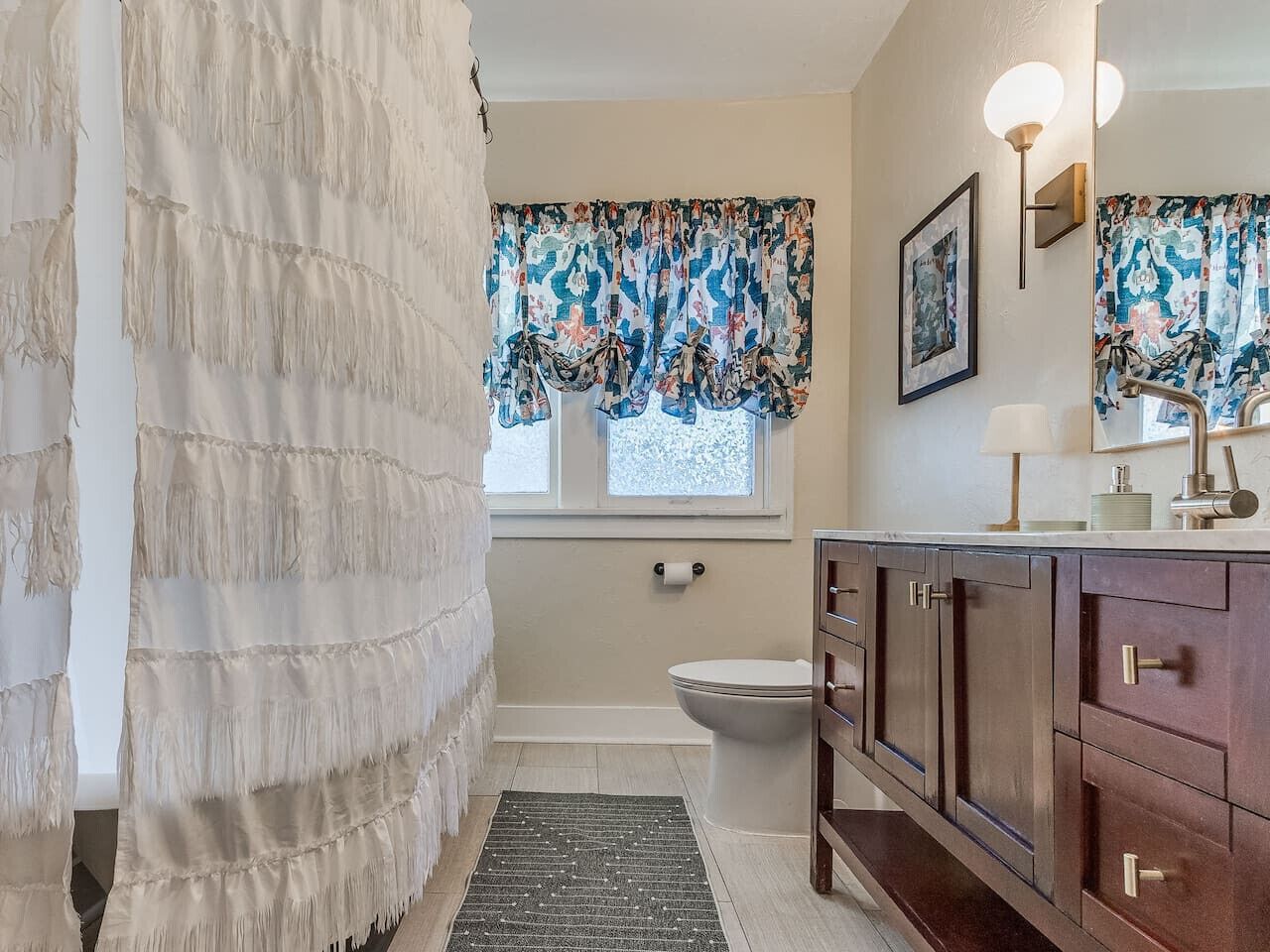Et badeværelse med toilet, vask med spejl, bruseforhæng og et vindue med blomstrede gardiner.