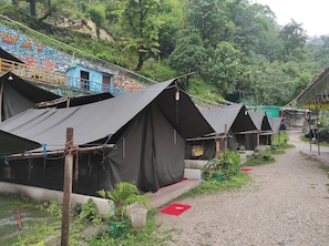 A row of black tents with a gravel path leading to them, surrounded by greenery and a mural on a building in the background.