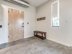 Ein Zimmer mit Holztür, einer Bank, einem kleinen Tisch, einem Fenster und einem an der Wand befestigten Regal.