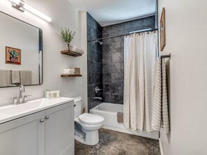 Un baño moderno con lavamanos, inodoro y bañera blancos. Hay una cortina de ducha, un espejo y un estante con toallas.