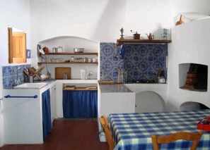 Eine kleine KĂŒche mit SpĂŒle, Herd und Regalen, ein Essbereich mit kariertem Tisch und ein kleines Fenster.