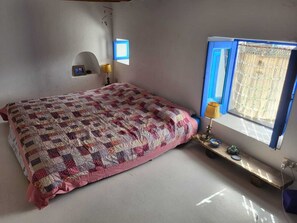 Ein Schlafzimmer mit einem Bett, einem Nachttisch, einer Lampe, einem Fenster mit blauen FensterlÀden und einer kleinen Ablage mit Dekoration.