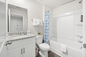 Et badeværelse med et hvidt toilet, en vask med en granitbordplade, en bruser med et hvidt gardin og et spejl over vasken.