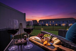 Una mesa con copas de vino, queso y frutas en un balcón con vista a edificios al anochecer.