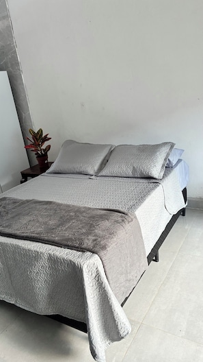 Ein Bett mit grauer, strukturierter Decke und Kissen, eine kleine Topfpflanze und eine schlichte weiße Wand.
