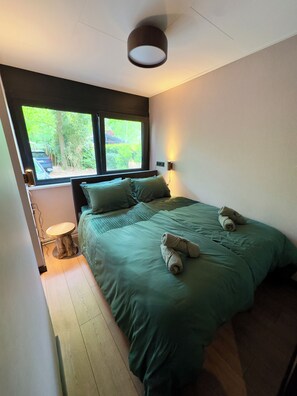 A bedroom with a bed, pillows, a bedside table, a lamp, and a window with a view of greenery. - New: Luxury forest house in nature with private hot tub (Rheezerveen)
