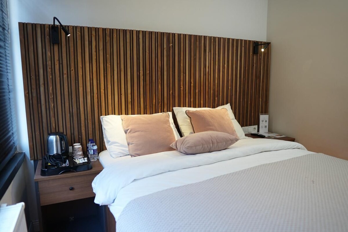 Habitación de hotel con cabecera de madera, cama con ropa blanca, dos almohadas, mesita de noche con cafetera, una botella de agua y una lámpara.