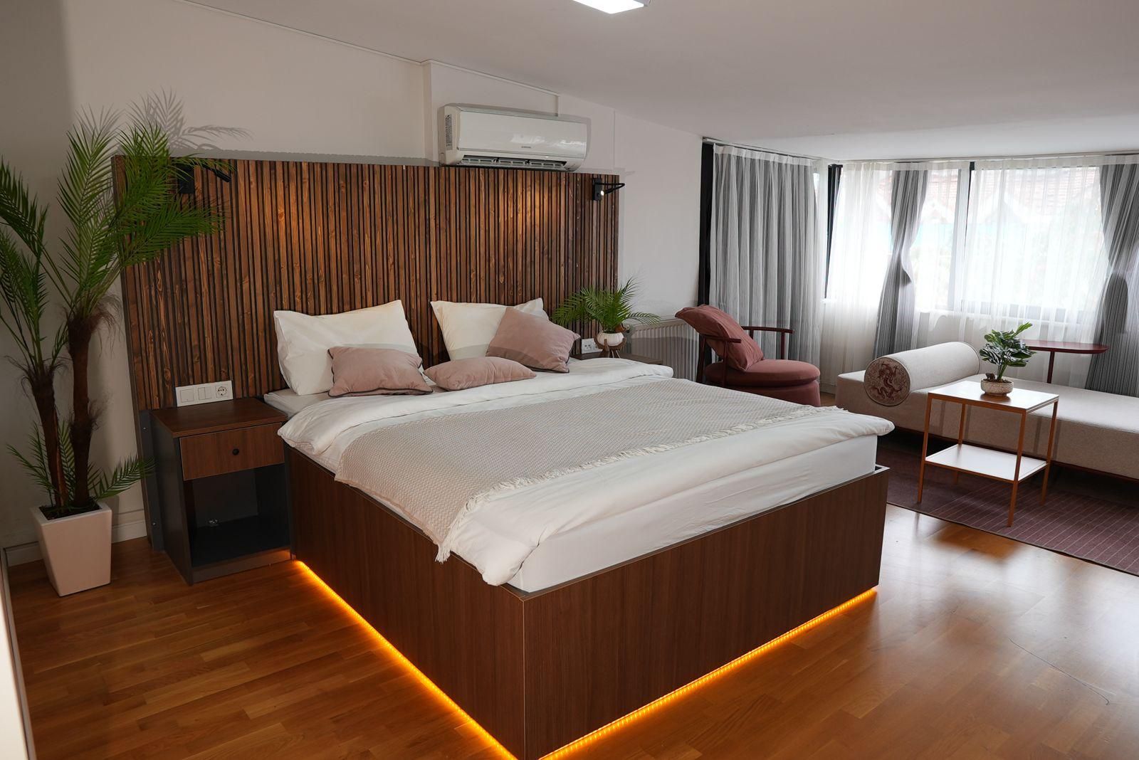 A modern bedroom with a large bed, bedside tables, a nightstand, a potted plant, a chair, a sofa, a small table, and a window with curtains.